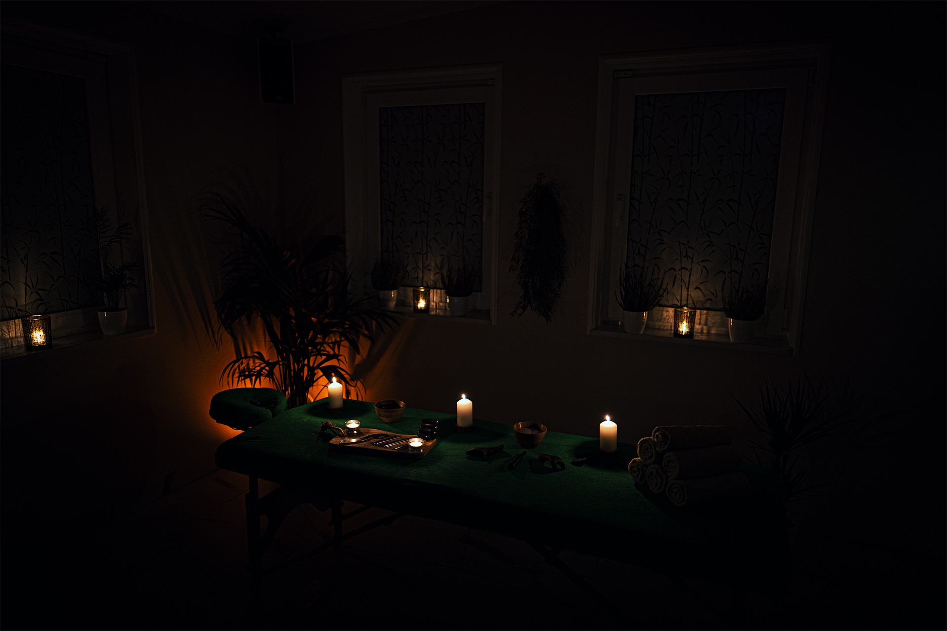 Przyciemnione wnętrze gabinetu z zielonym łóżkiem do masażu, świecami i roślinami doniczkowymi w oknach.