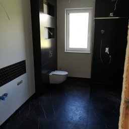 DEKOR PAWEŁ SKRZELIŃSKI - Częściowo wykończona łazienka z czarnymi płytkami imitującymi marmur na podłodze i ścianach, biała toaleta, kabina prysznicowa z czarną ścianą i wystające kable sufitowe.