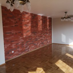 DEKOR PAWEŁ SKRZELIŃSKI - Salon z parkietem w jodełkę i ścianą z cegły klinkierowej, oświetlony przez wiszące lampy i promienie słoneczne wpadające przez okno.