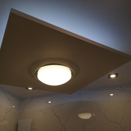 DEKOR PAWEŁ SKRZELIŃSKI - Sufit podwieszany z centralnym okrągłym oświetleniem i dodatkowymi punktami świetlnymi w wykończonym wnętrzu z marmurowymi płytkami na ścianach.