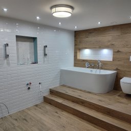 DEKOR PAWEŁ SKRZELIŃSKI - Nowoczesna łazienka w trakcie wykańczania, z białą wolnostojącą wanną na drewnianym podeście, częściowo wyłożona białymi płytkami imitującymi cegłę i drewnopodobnymi panelami ściennymi, widoczne...