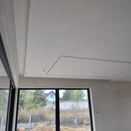 DEKOR PAWEŁ SKRZELIŃSKI - Surowy stan wnętrza z dużymi oknami, widoczne ściany po nałożeniu warstwy wygładzającej, podwieszany sufit z otworami na oświetlenie, kable zwisające z sufitu.