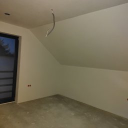 DEKOR PAWEŁ SKRZELIŃSKI - Surowe wnętrze poddasza ze ścianami po gładziowaniu, widoczne wystające kable z sufitu, przygotowane do dalszych prac wykończeniowych.