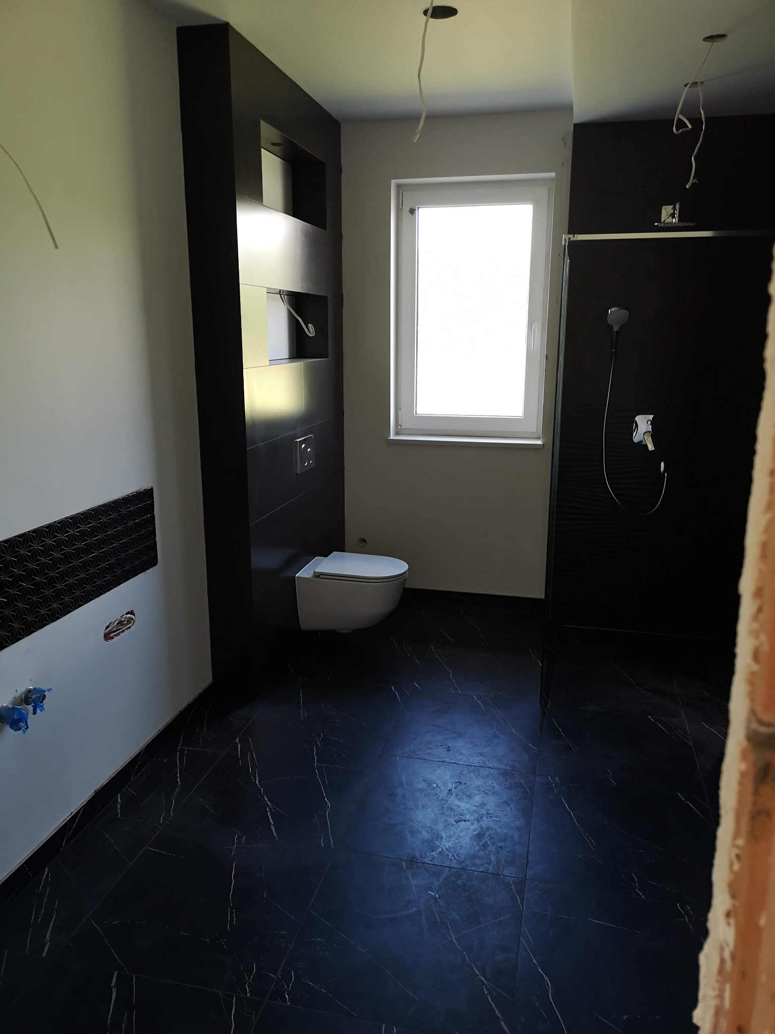 Częściowo wykończona łazienka z czarnymi płytkami imitującymi marmur na podłodze i ścianach, biała toaleta, kabina prysznicowa z czarną ścianą i wystające kable sufitowe.