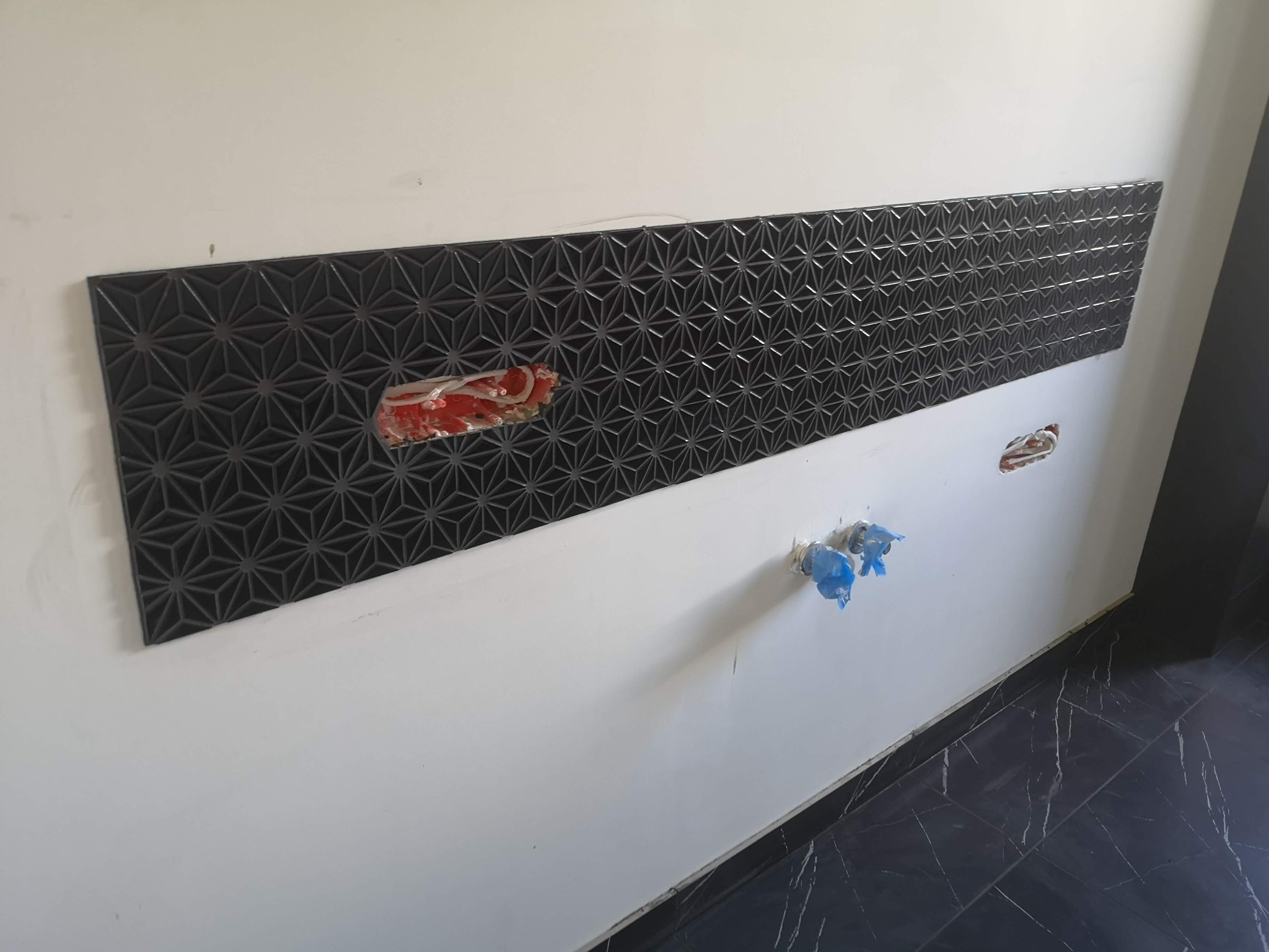 Fragment ściany z zamontowaną dekoracyjną, czarną płytą o geometrycznym wzorze gwiazd. Widoczne niezabezpieczone kable i rury, przygotowane do podłączenia armatury.