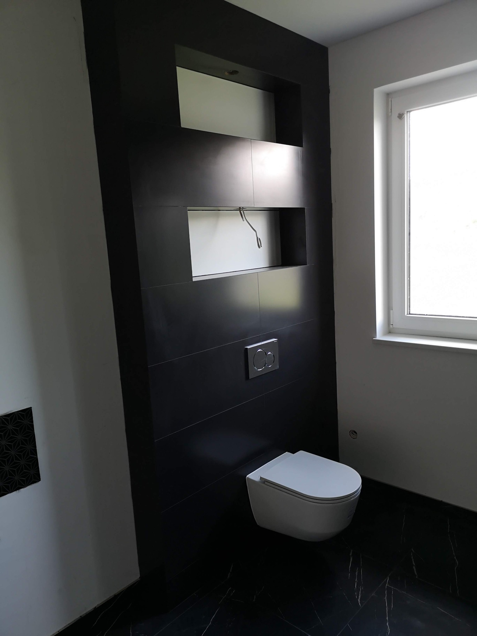 Nowoczesna toaleta podwieszana na czarnej ścianie z wnękami, marmurowa podłoga, widoczne przyłącza w górnej wnęce.
