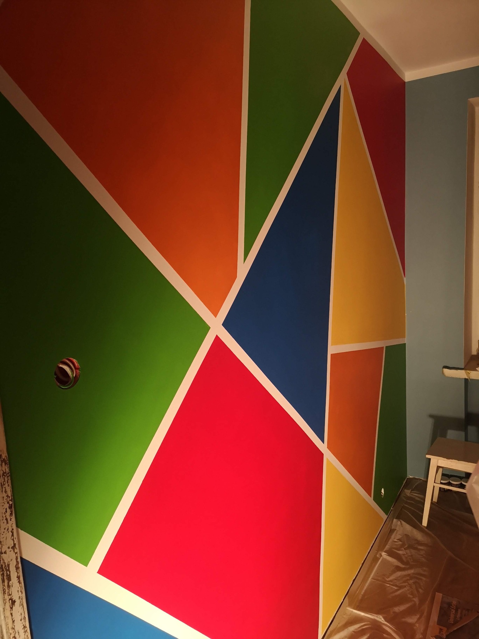 Ściana pomalowana w geometryczne wzory z użyciem taśmy malarskiej, widoczna puszka elektryczna i zabezpieczona folią podłoga.