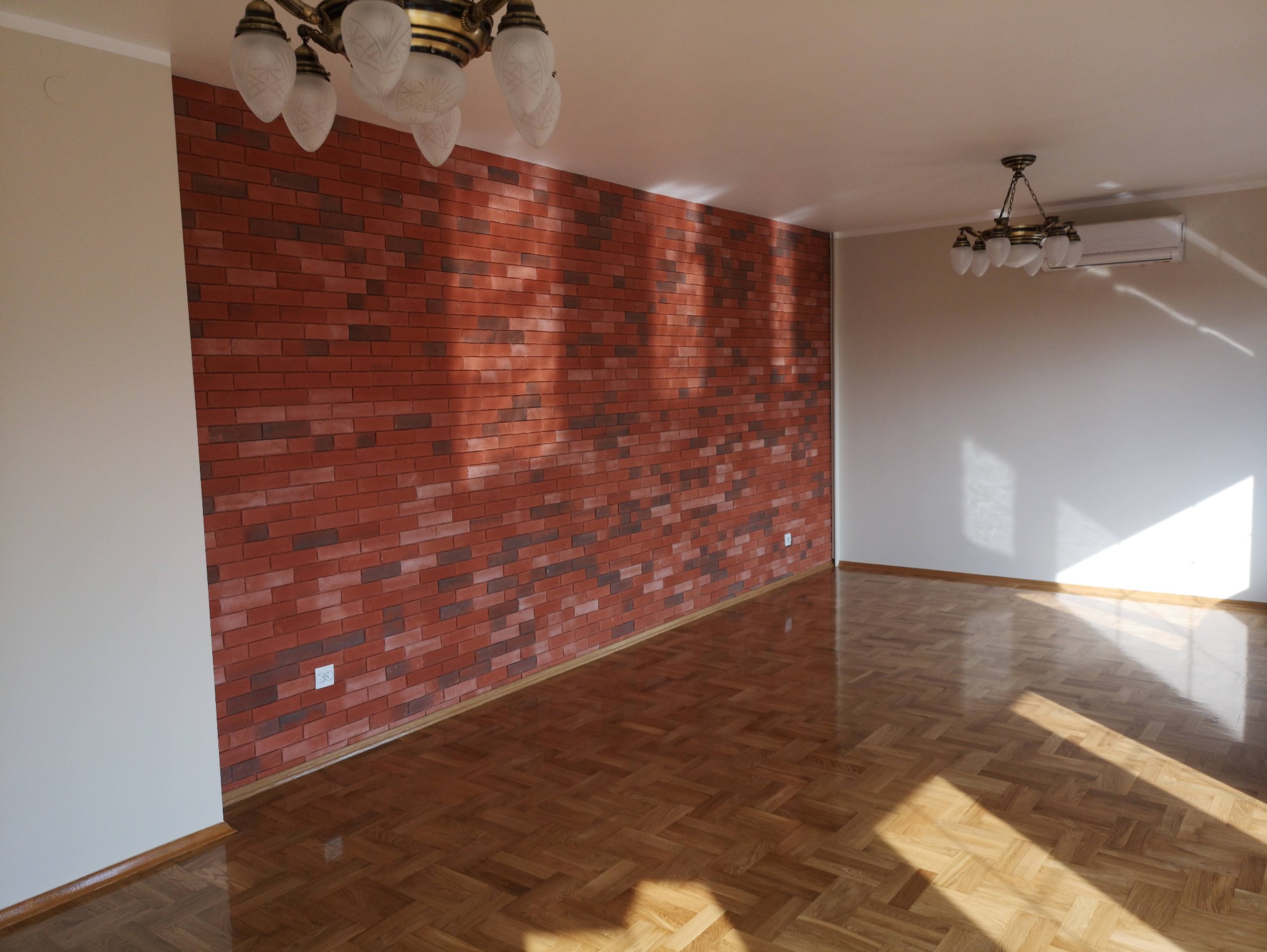 Salon z parkietem w jodełkę i ścianą z cegły klinkierowej, oświetlony przez wiszące lampy i promienie słoneczne wpadające przez okno.