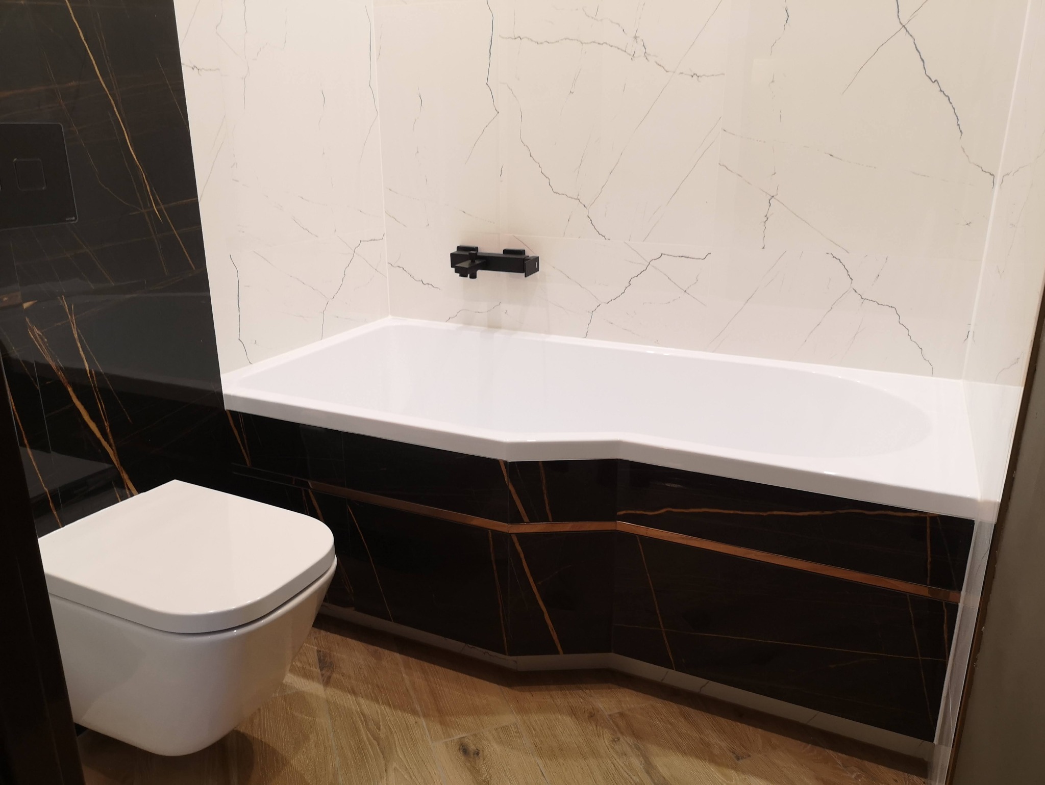 Nowoczesna łazienka z czarno-białymi marmurkowymi płytkami, białą wanną i podwieszaną toaletą, minimalistyczna armatura w kolorze czarnym.