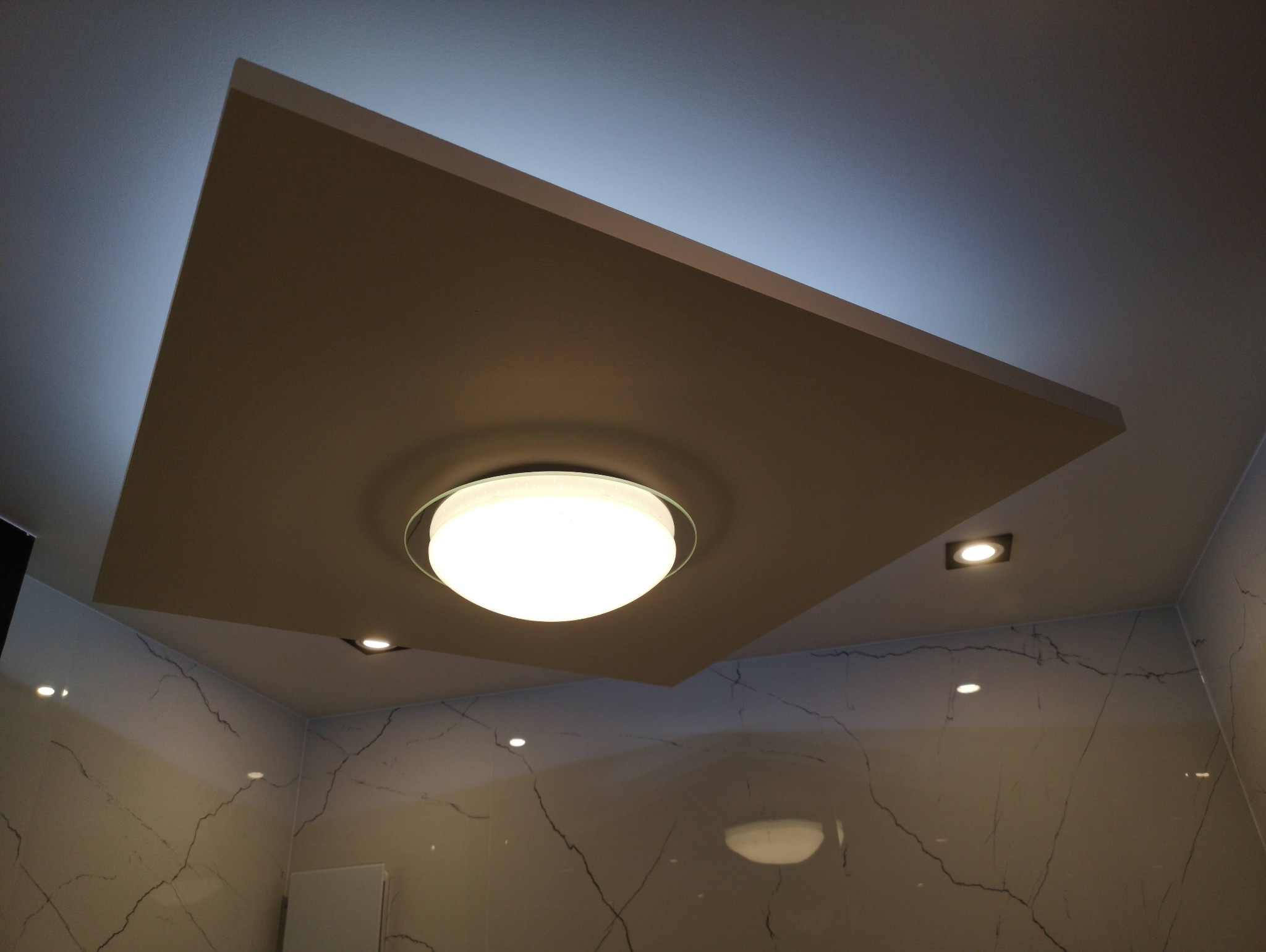 Sufit podwieszany z centralnym okrągłym oświetleniem i dodatkowymi punktami świetlnymi w wykończonym wnętrzu z marmurowymi płytkami na ścianach.