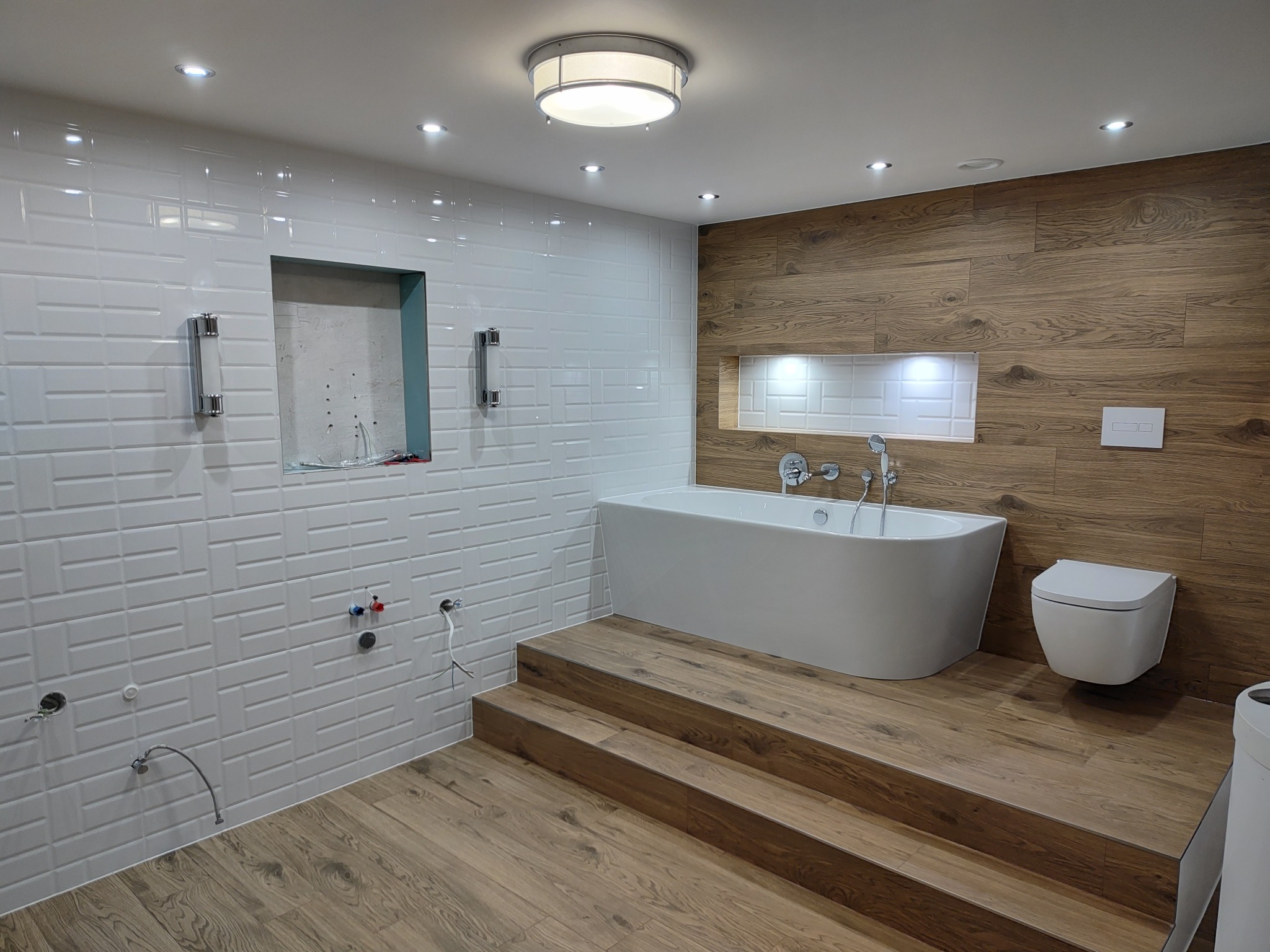 Nowoczesna łazienka w trakcie wykańczania, z białą wolnostojącą wanną na drewnianym podeście, częściowo wyłożona białymi płytkami imitującymi cegłę i drewnopodobnymi panelami ściennymi, widoczne...