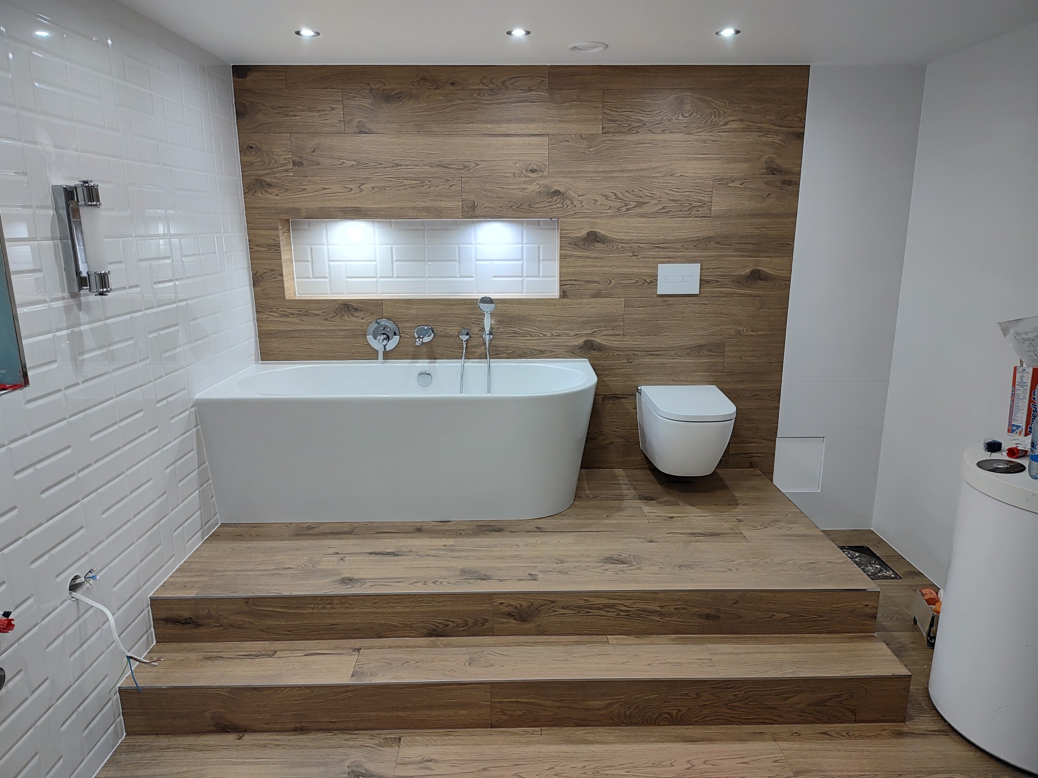 Nowoczesna łazienka z drewnopodobnymi płytkami na ścianach i podłodze, biała wanna wolnostojąca na podwyższeniu z trzema stopniami, wisząca toaleta i wnęka z oświetleniem.