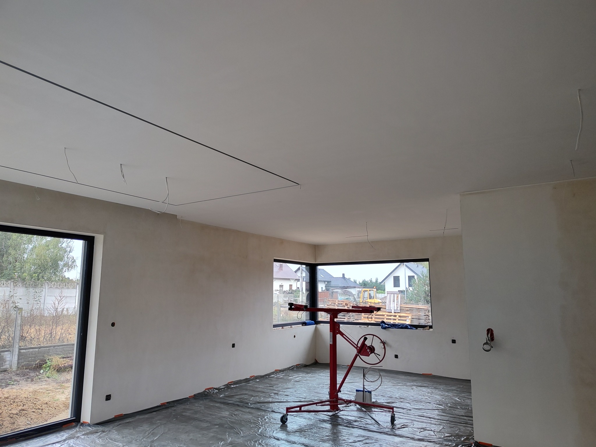 Wnętrze w trakcie prac wykończeniowych; widoczny podnośnik do płyt, ściany przygotowane pod malowanie, okna z czarnymi ramami, podłoga zabezpieczona folią.