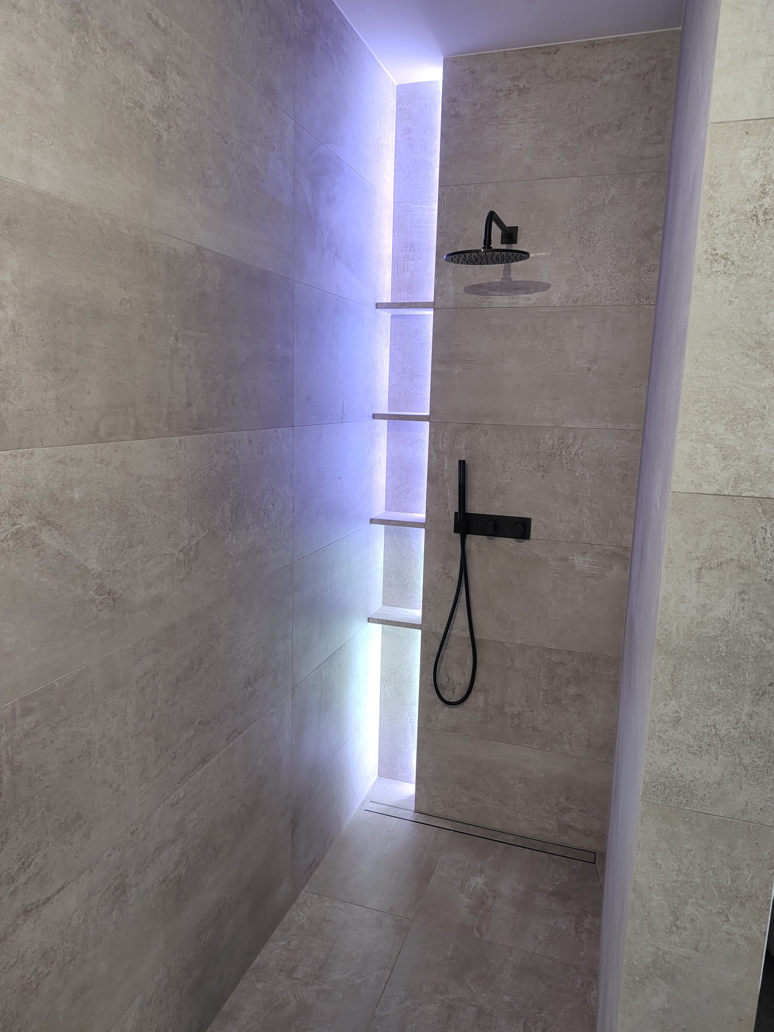 Nowoczesna kabina prysznicowa z beżowymi płytkami, czarną armaturą i podświetlanymi półkami w ścianie.