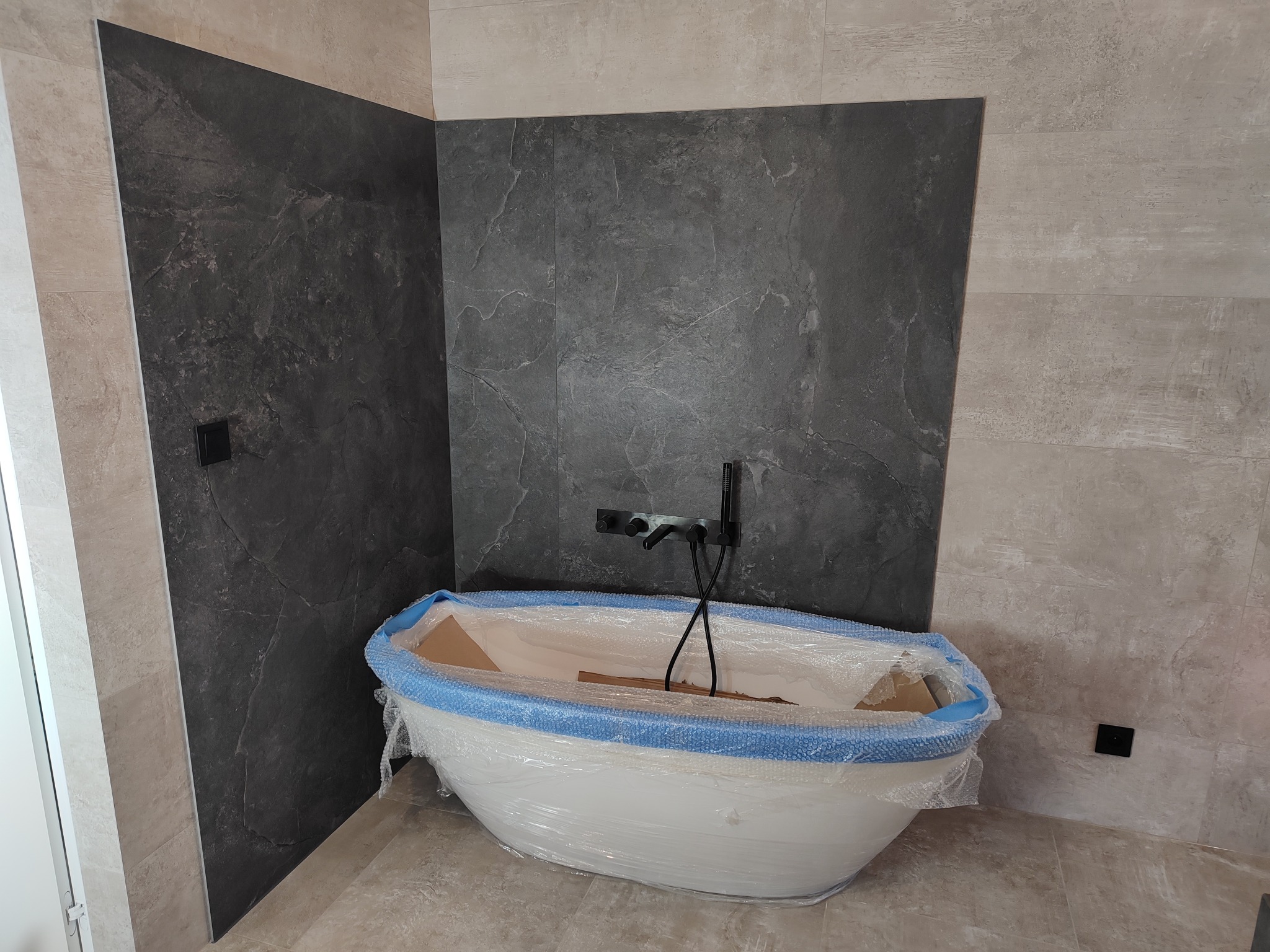 Wanna wolnostojąca zabezpieczona folią ochronną w łazience w Częstochowie, z ciemną armaturą na tle ściany wyłożonej płytami imitującymi kamień.