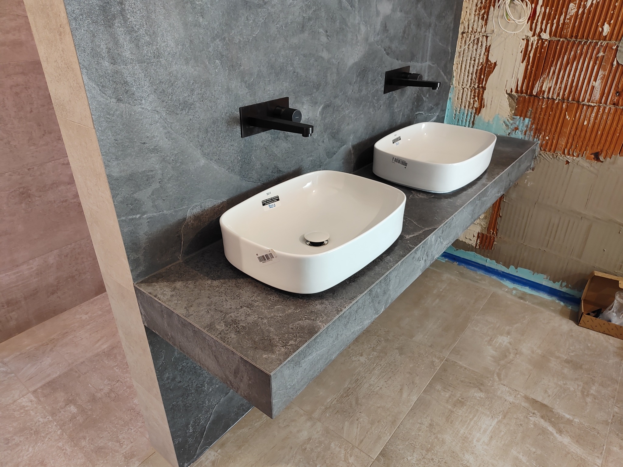 Dwie białe, prostokątne umywalki nablatowe z czarnymi bateriami zamontowane na szarym blacie z efektem kamienia, widoczne niedokończone fragmenty ściany z cegły i instalacji, podłoga wyłożona...