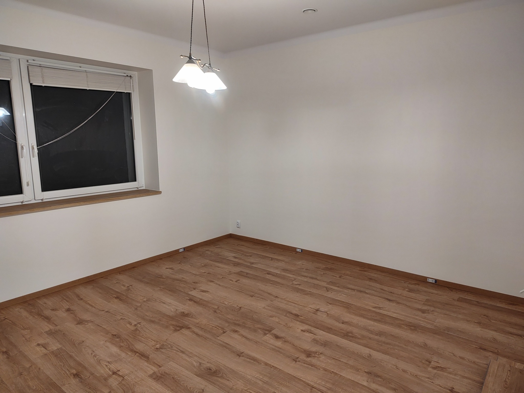 Puste pomieszczenie z panelami podłogowymi o barwie drewna, białymi ścianami, oknem z żaluzjami i lampą wiszącą z trzema kloszami.