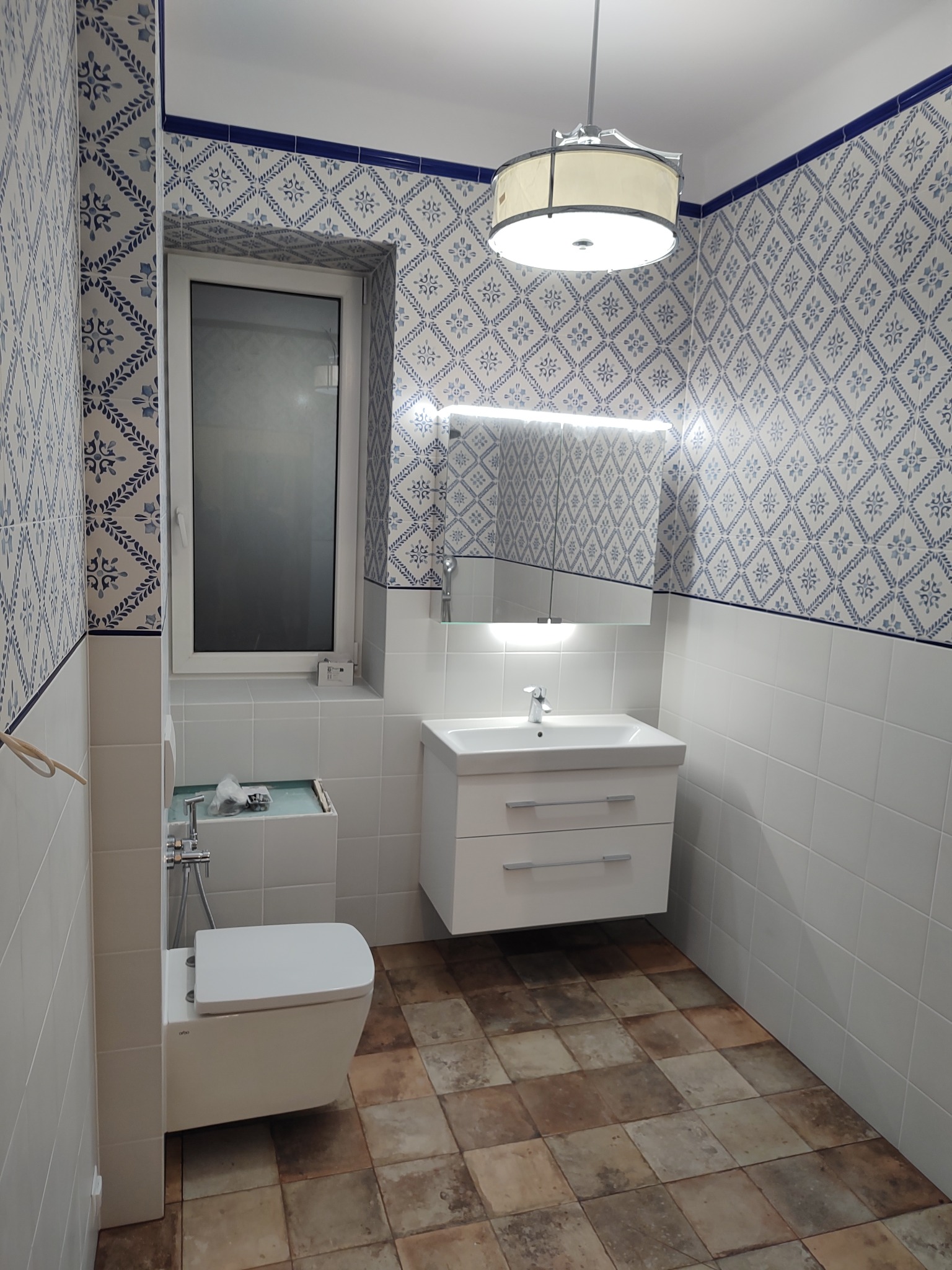 Nowoczesna łazienka z wzorzystymi, niebiesko-białymi płytkami na ścianach, białymi płytkami do połowy ściany, brązową podłogą z płytek, wiszącą szafką z umywalką i lustrem z oświetleniem LED,...