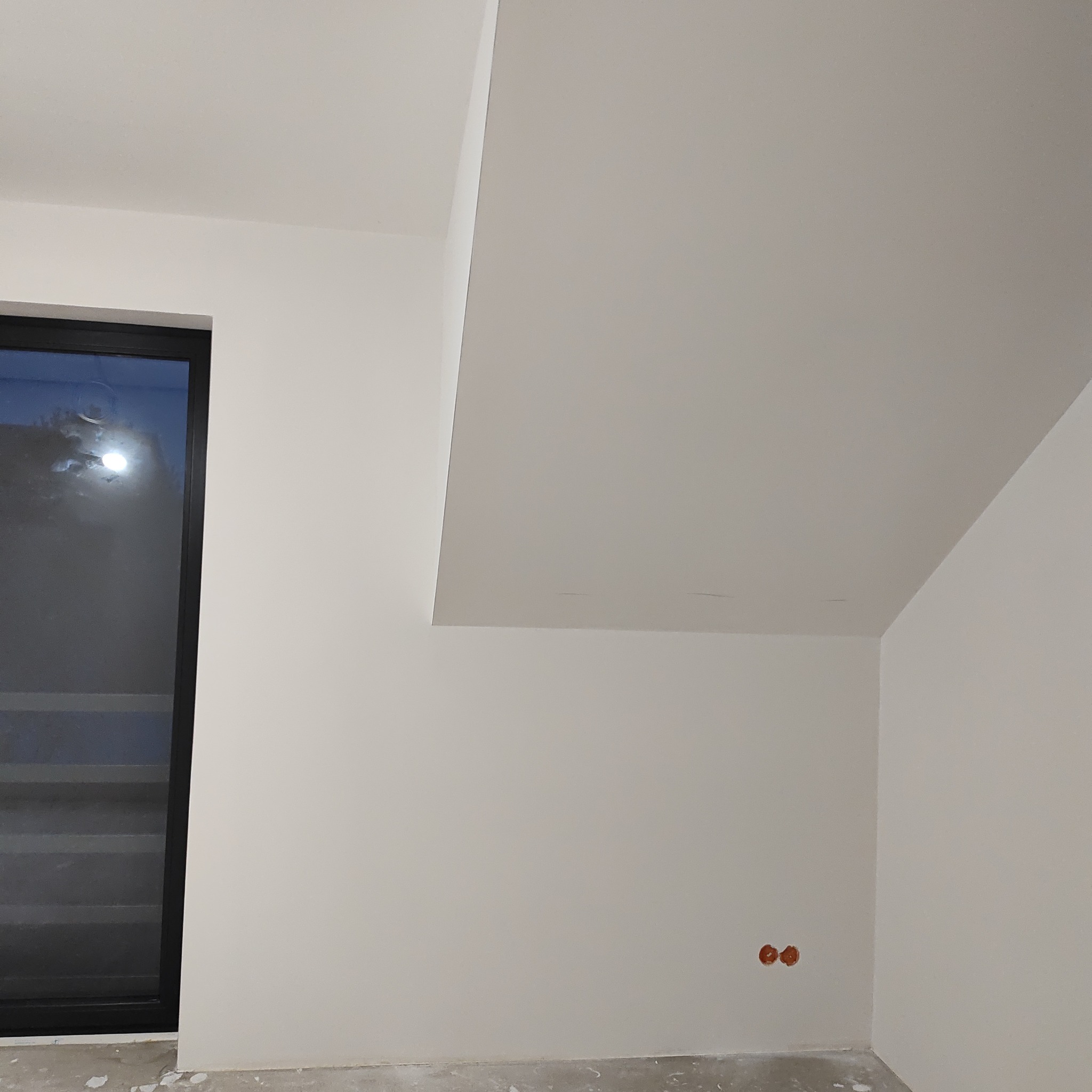 Wnętrze pokoju po świeżym malowaniu na biało, widoczne gniazdka elektryczne i fragment okna z czarną ramą, na podłodze resztki po remoncie.