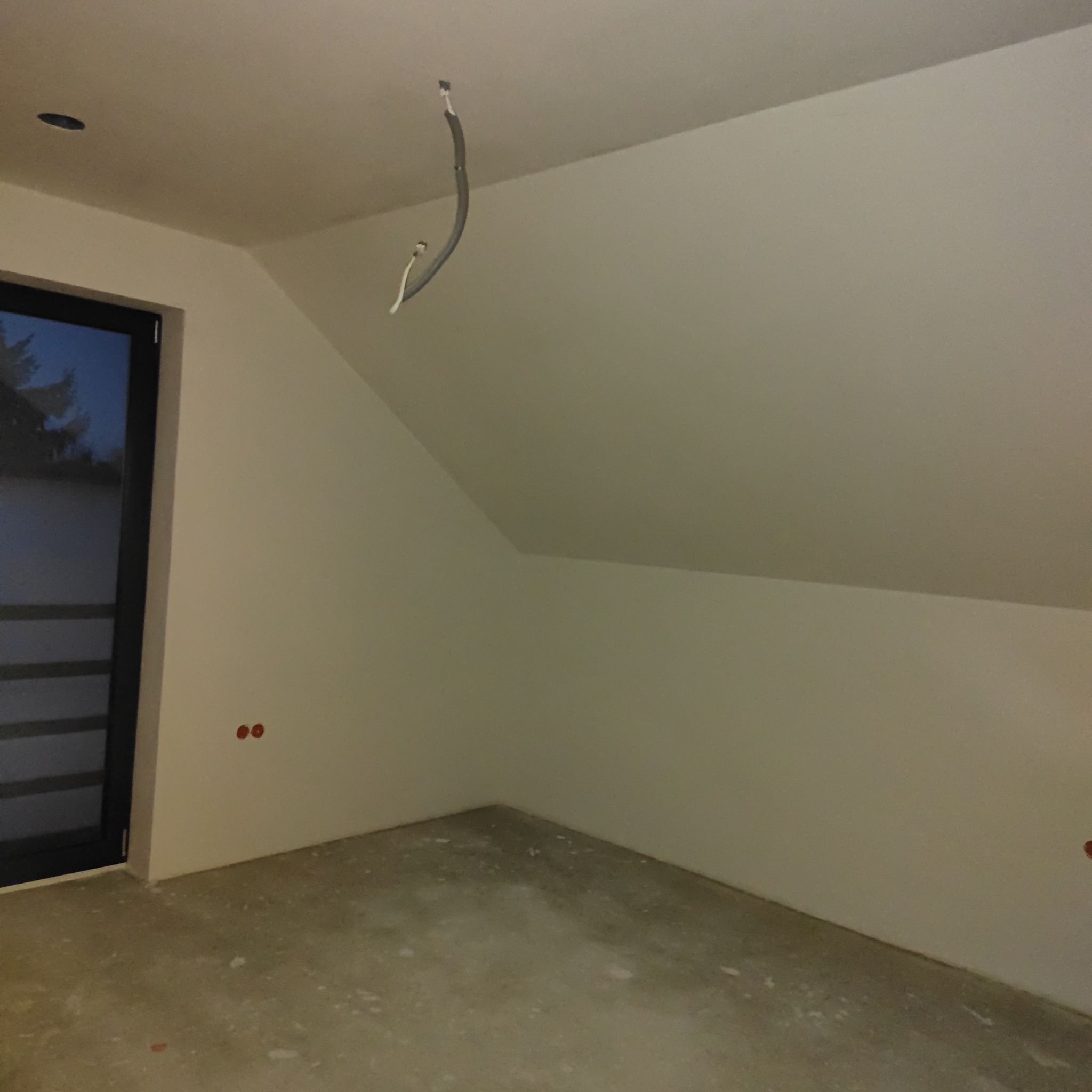 Surowe wnętrze poddasza ze ścianami po gładziowaniu, widoczne wystające kable z sufitu, przygotowane do dalszych prac wykończeniowych.