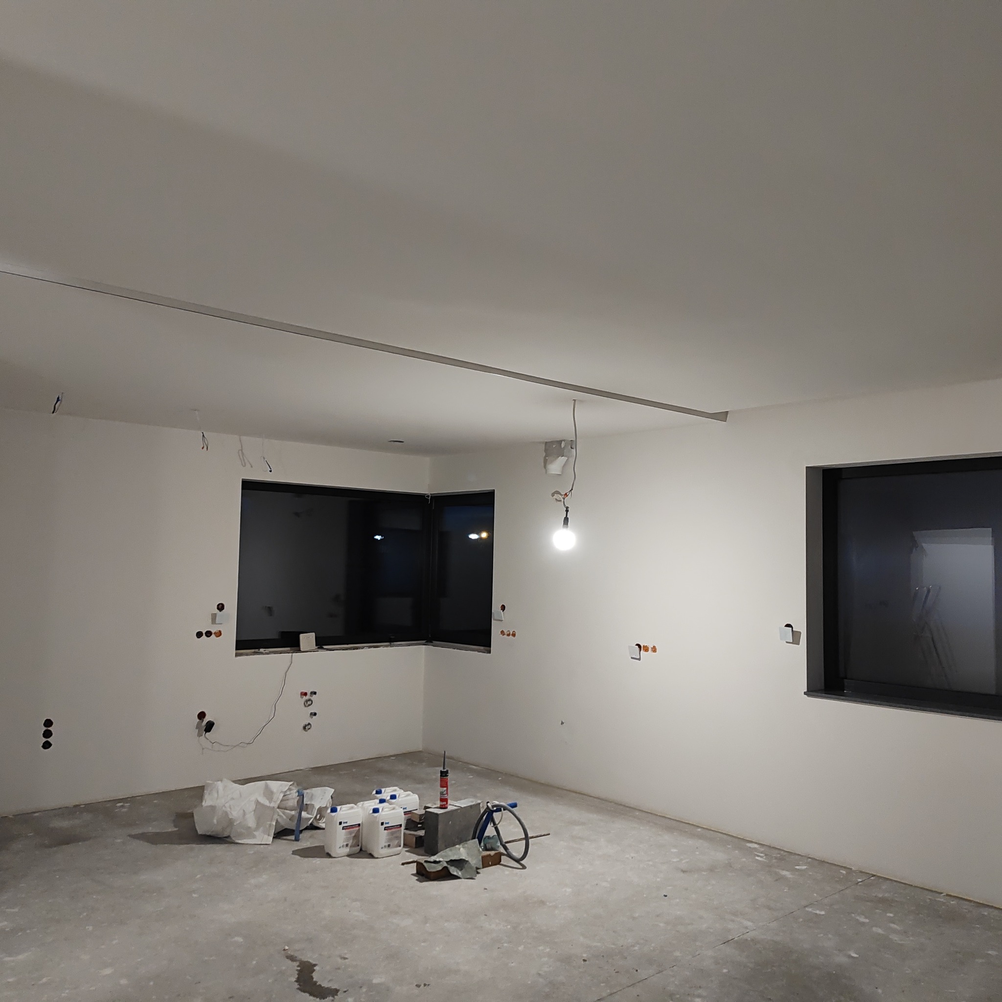 Pokój w trakcie prac wykończeniowych; ściany wyszpachlowane, widoczne puszki elektryczne, podłoga surowa, przygotowane materiały budowlane i narzędzia.