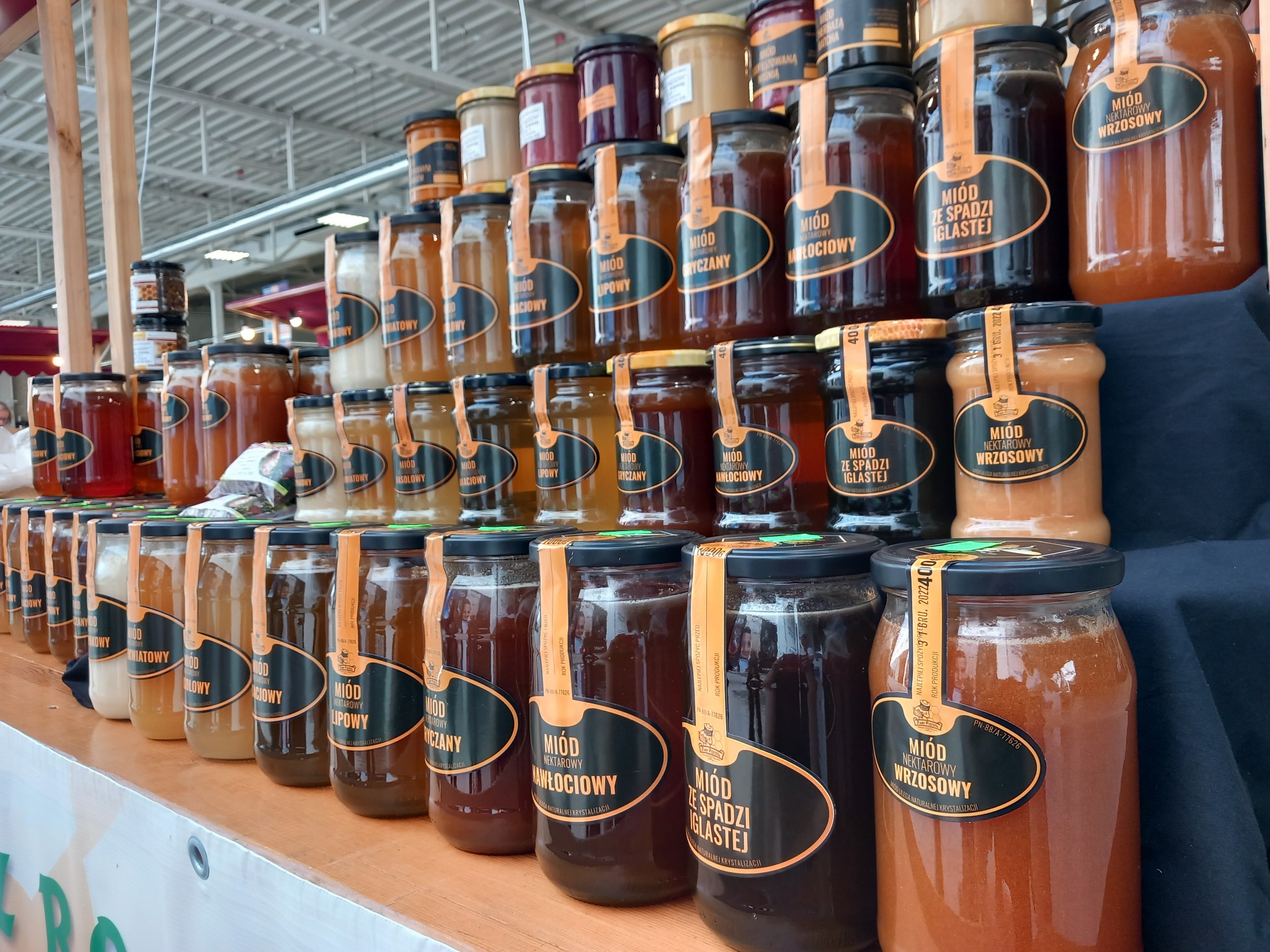 Rzędy słoików z różnymi rodzajami miodu ustawione na półkach, widoczne etykiety informujące o rodzaju miodu, od jasnych do ciemnych odcieni, z metalowymi zakrętkami.