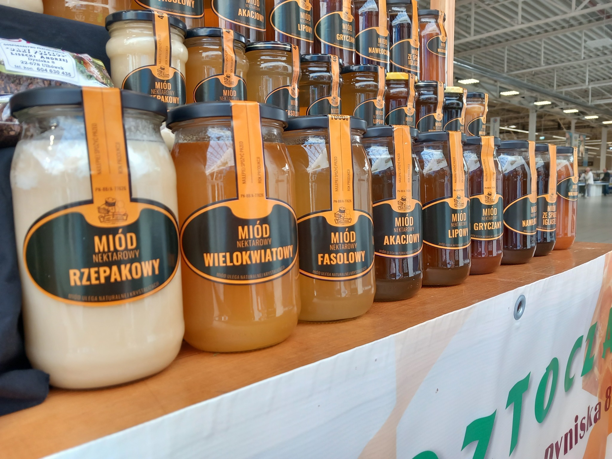 Rząd słoików z różnymi rodzajami miodu (rzepakowy, wielokwiatowy, fasolowy, akacjowy, lipowy, gryczany, nawłociowy, ze spadzi iglastej) na drewnianej półce, widoczne etykiety z nazwami...