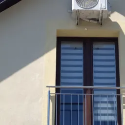 Biała jednostka zewnętrzna klimatyzatora marki Gree zamontowana na elewacji budynku nad oknem balkonowym z żaluzjami poziomymi.