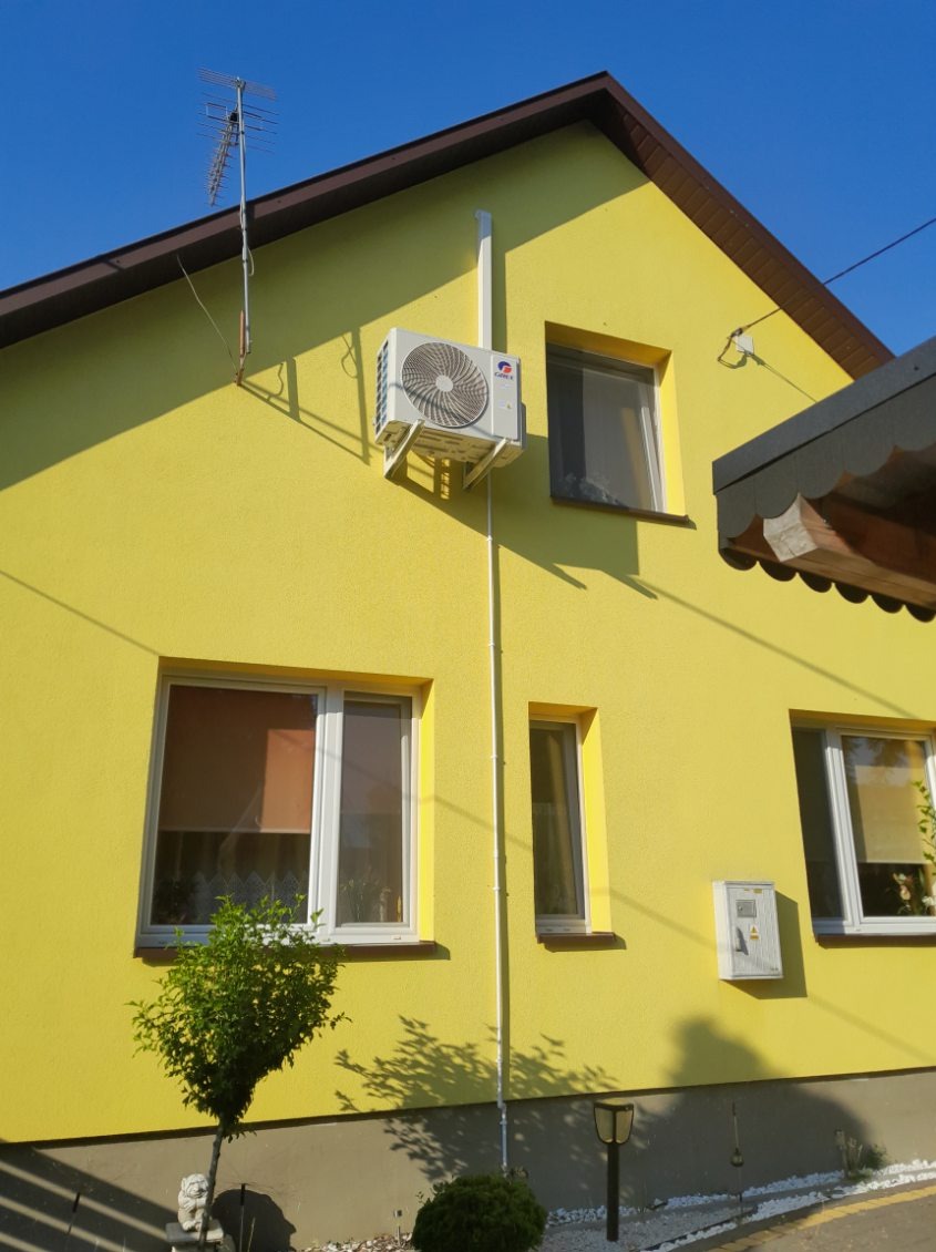 Żółty dom jednorodzinny z zamontowaną jednostką zewnętrzną klimatyzacji, widoczne okna i antena telewizyjna na dachu.