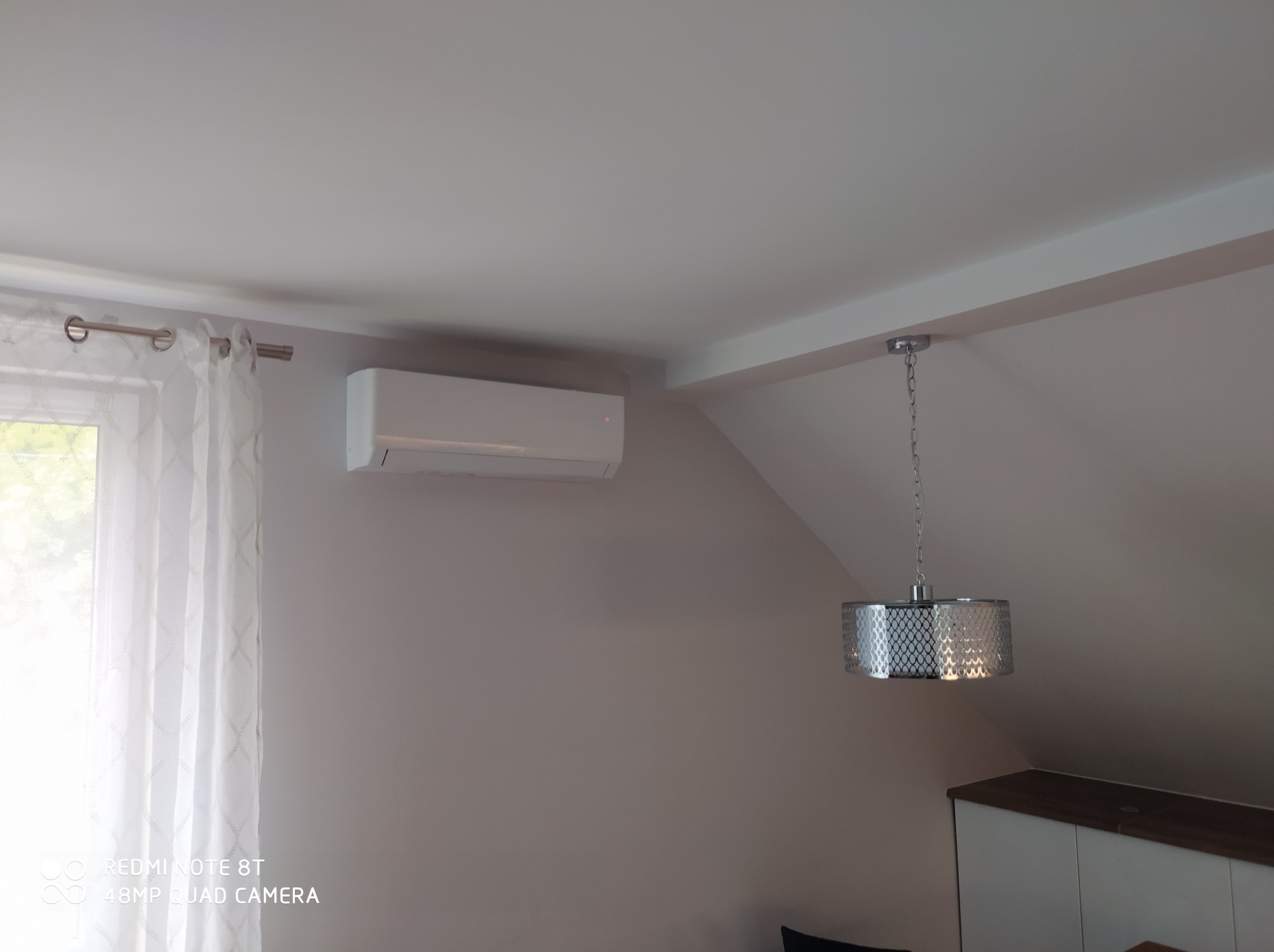 Biały klimatyzator ścienny zamontowany w pokoju na poddaszu, obok okna z firanką i ozdobnej lampy wiszącej.