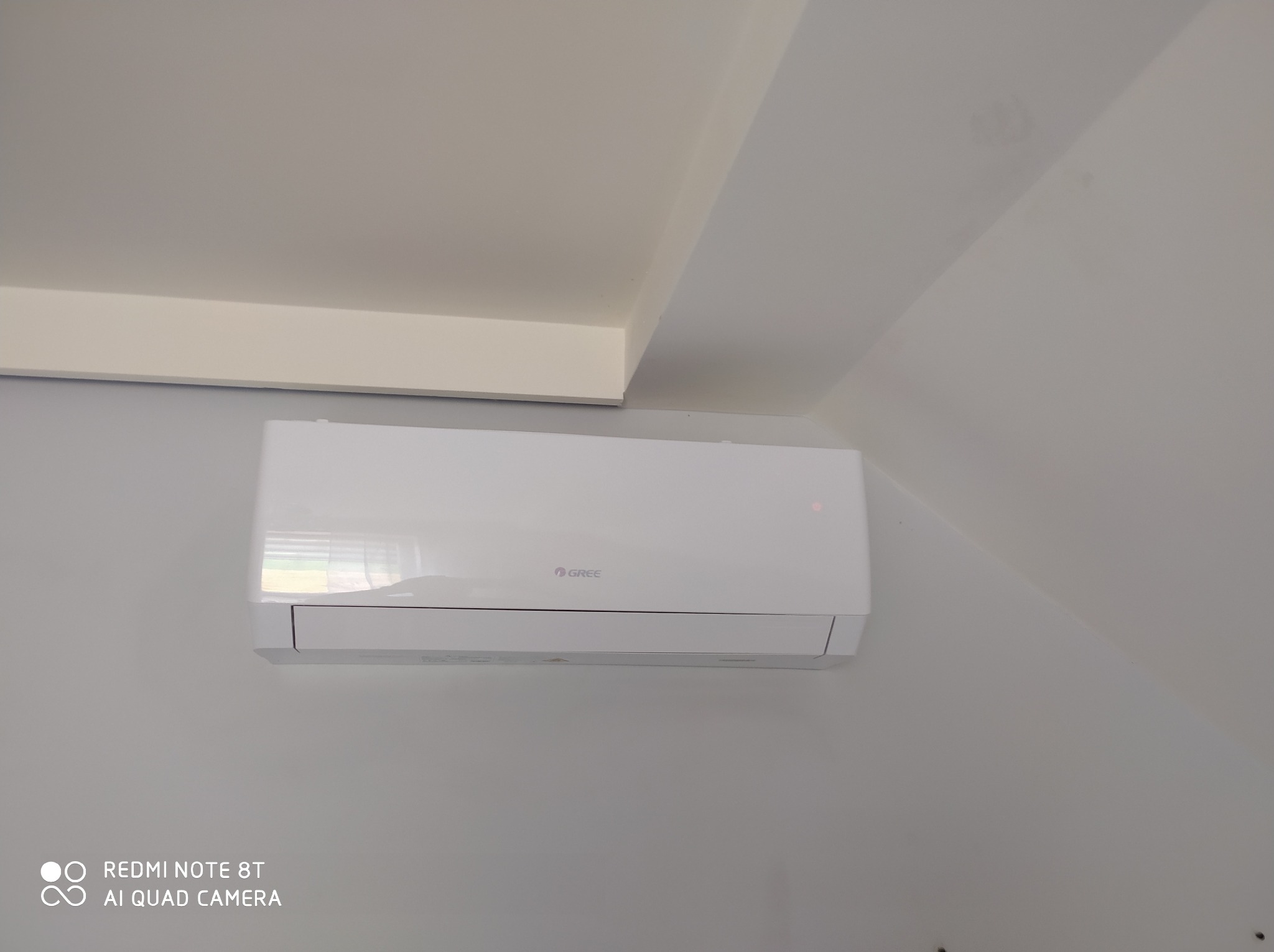 Biały klimatyzator marki Gree zamontowany na jasnej ścianie w pomieszczeniu o skośnym suficie, widoczny z bliska.