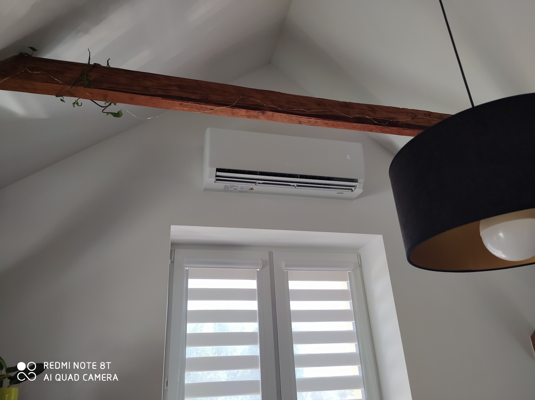 Biały klimatyzator ścienny zamontowany nad oknem z żaluzjami w pokoju na poddaszu, widoczna drewniana belka stropowa z bluszczem i fragment czarnej lampy.