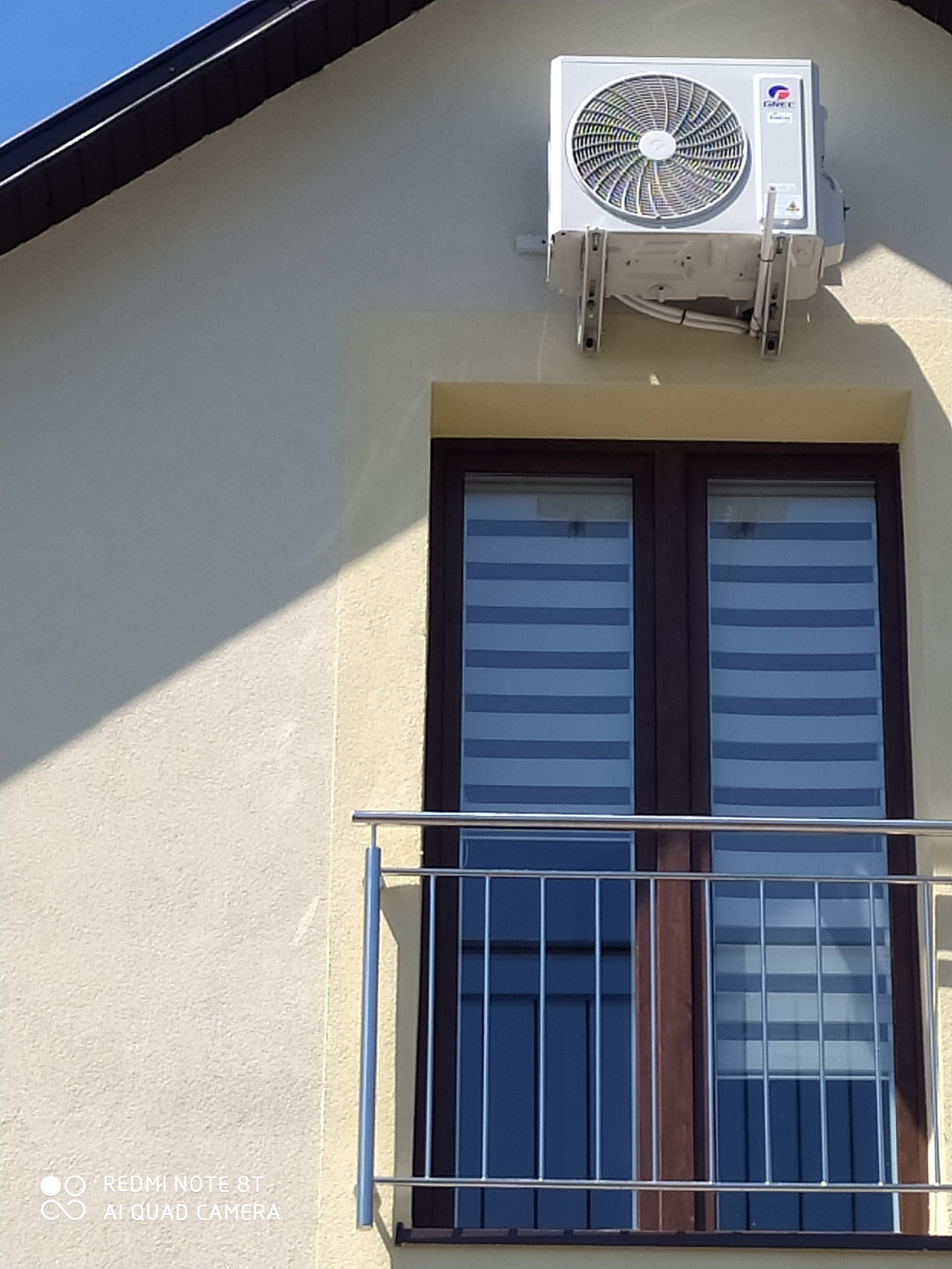 Biała jednostka zewnętrzna klimatyzatora marki Gree zamontowana na elewacji budynku nad oknem balkonowym z żaluzjami poziomymi.