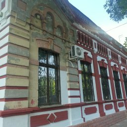 Saks Bud spolka z ograniczona odpowiedzialnoscia - Fragment odnowionej elewacji budynku z czerwono-białymi akcentami, widoczne okna z ozdobnymi kratami, klimatyzator na ścianie i ślady graffiti w dolnej części.