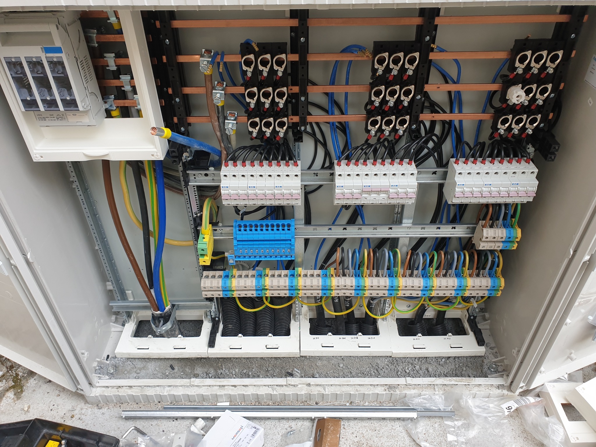 Otwarta skrzynka rozdzielcza z widocznymi bezpiecznikami, listwami zaciskowymi, przewodami w różnych kolorach i miedzianymi szynami zbiorczymi, zamontowana na szarej, betonowej powierzchni.