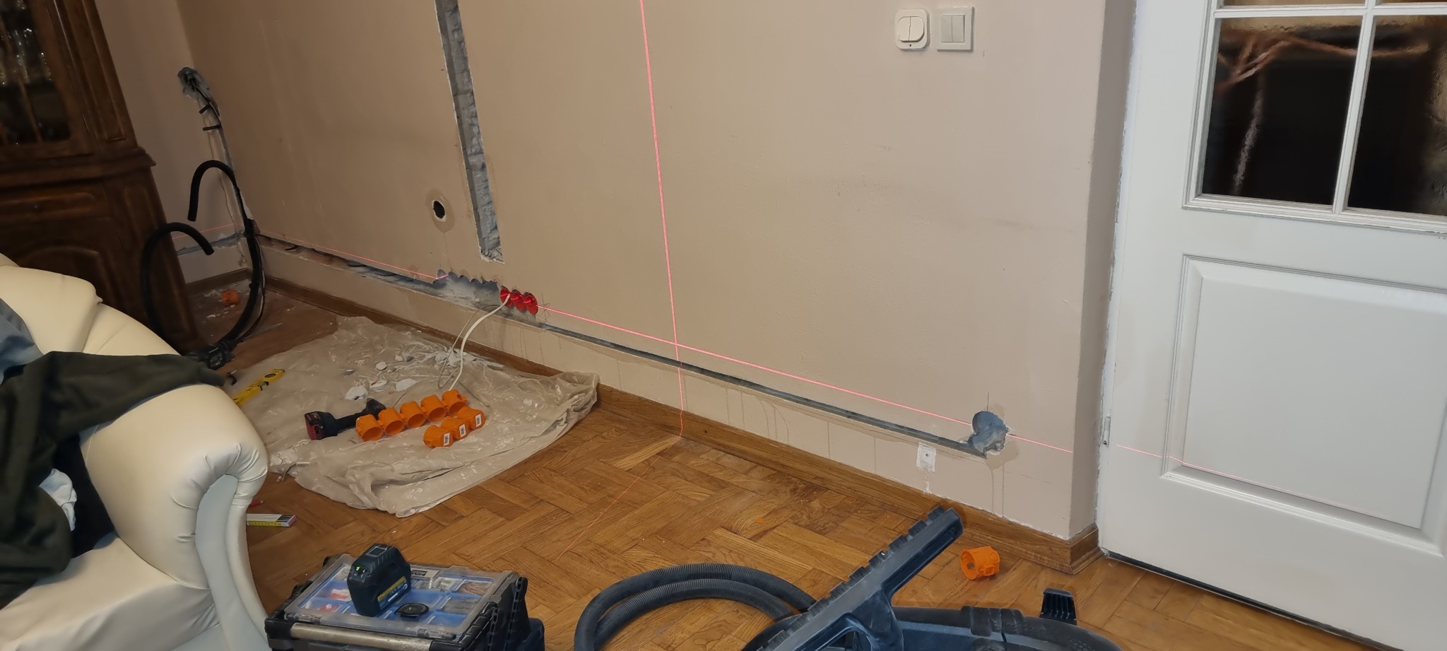 Instalacja elektryczna w trakcie remontu: wyżłobiona ściana z widocznymi przewodami, czerwone puszki elektryczne, poziomica laserowa wyznaczająca linię, narzędzia i osprzęt na podłodze...