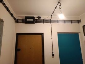 Widok na korytarz z widoczną, natynkową instalacją elektryczną w czarnych peszlach, prowadzoną wzdłuż sufitu i ściany, z zamontowaną skrzynką bezpiecznikową i pojedynczą lampą sufitową na długim...