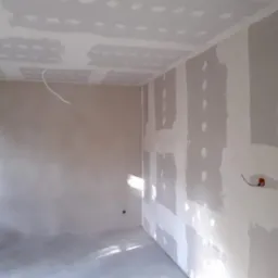 Pokój w trakcie remontu: ściany i sufit pokryte płytami gipsowo-kartonowymi, widoczne łączenia wymagające szpachlowania, wystające przewody elektryczne, częściowo wygładzona ściana w tle, światło...