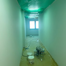 Mateusz Adamczewski - Korytarz w trakcie malowania natryskowego na jasnozielono, z zabezpieczoną folią podłogą i sufitem, widoczne wiadra z farbą i urządzenie malarskie.