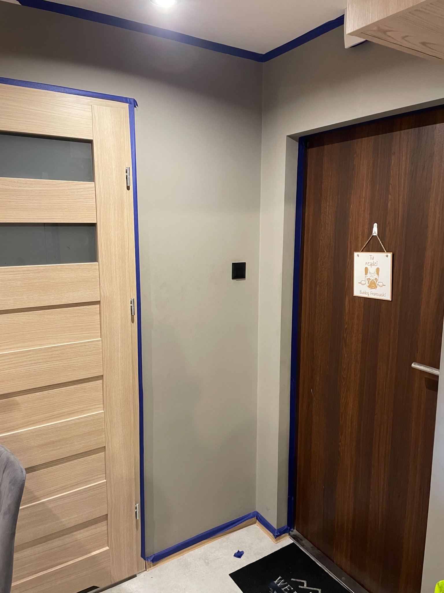 Przygotowanie do malowania ścian w przedpokoju: ściany zabezpieczone niebieską taśmą malarską wokół drzwi drewnianych i ościeżnic, widoczny fragment sufitu i podłogi.
