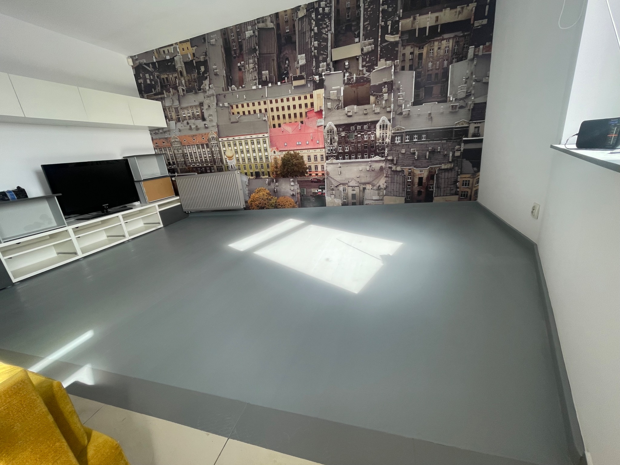 Szare, jednolite wykończenie podłogi w pokoju z fototapetą przedstawiającą panoramę miasta, widoczny fragment żółtego fotela.