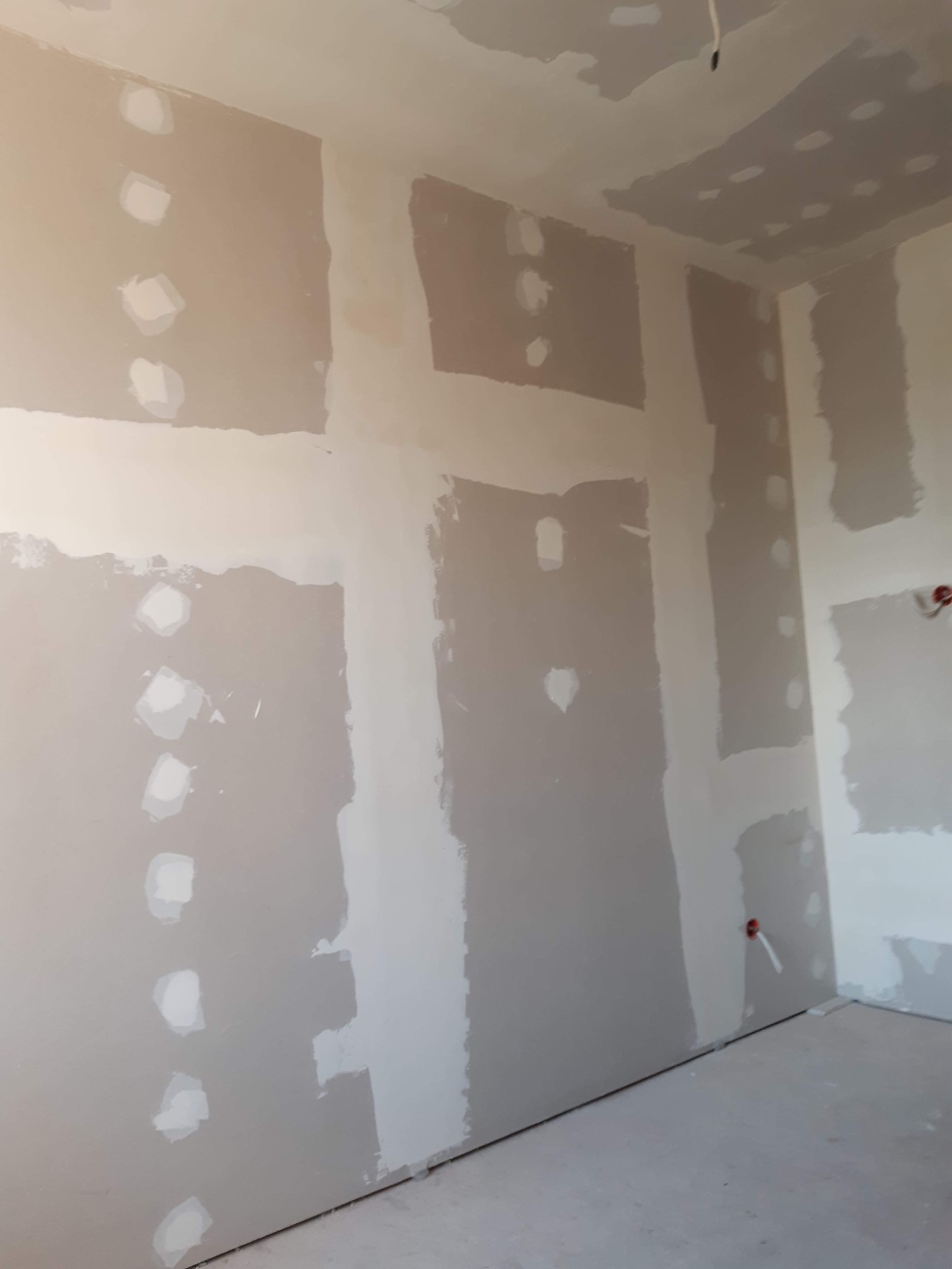 Wnętrze pokoju w trakcie prac wykończeniowych, widoczne ściany z płyt kartonowo-gipsowych ze szpachlowanymi łączeniami i otworami po wkrętach, przygotowane do dalszych prac.