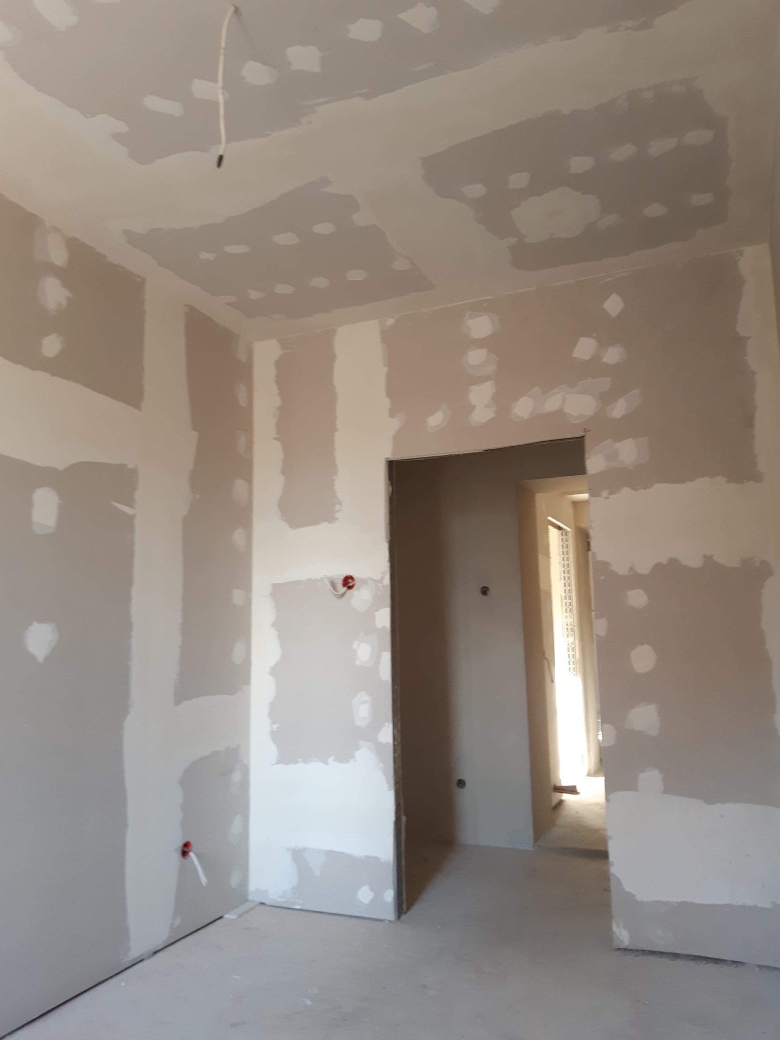 Pokój w trakcie prac wykończeniowych, widoczne płyty kartonowo-gipsowe na ścianach i suficie, częściowo pokryte szpachlą, wystające kable elektryczne.