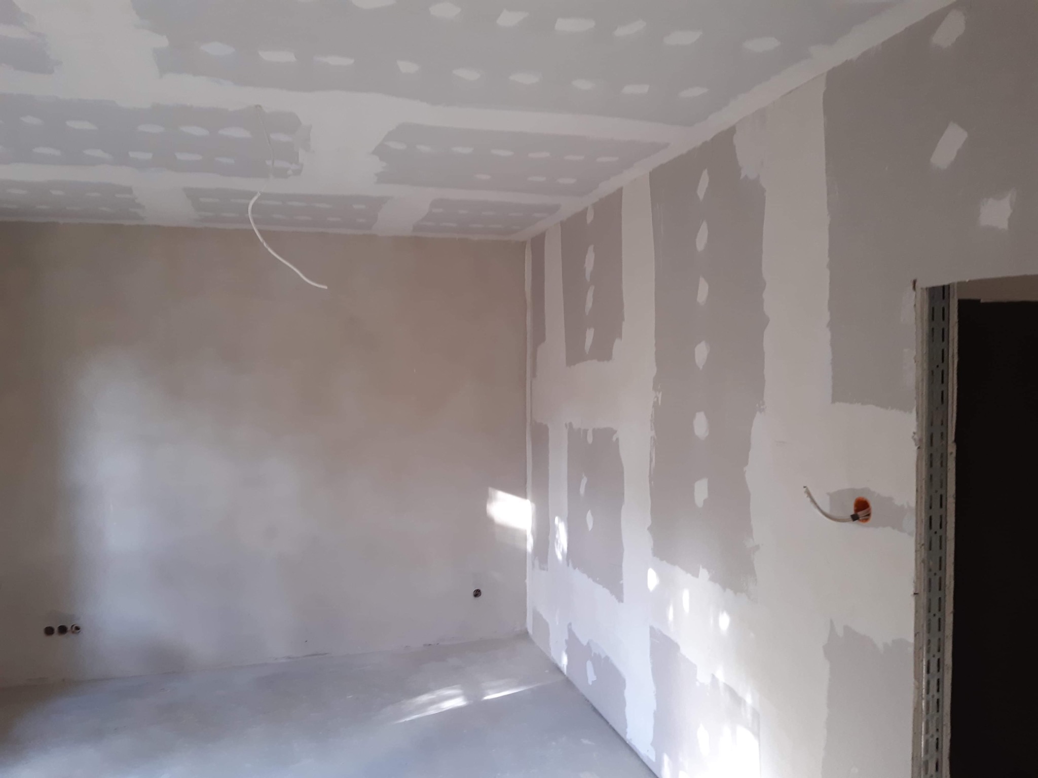 Pokój w trakcie remontu: ściany i sufit pokryte płytami gipsowo-kartonowymi, widoczne łączenia wymagające szpachlowania, wystające przewody elektryczne, częściowo wygładzona ściana w tle, światło...