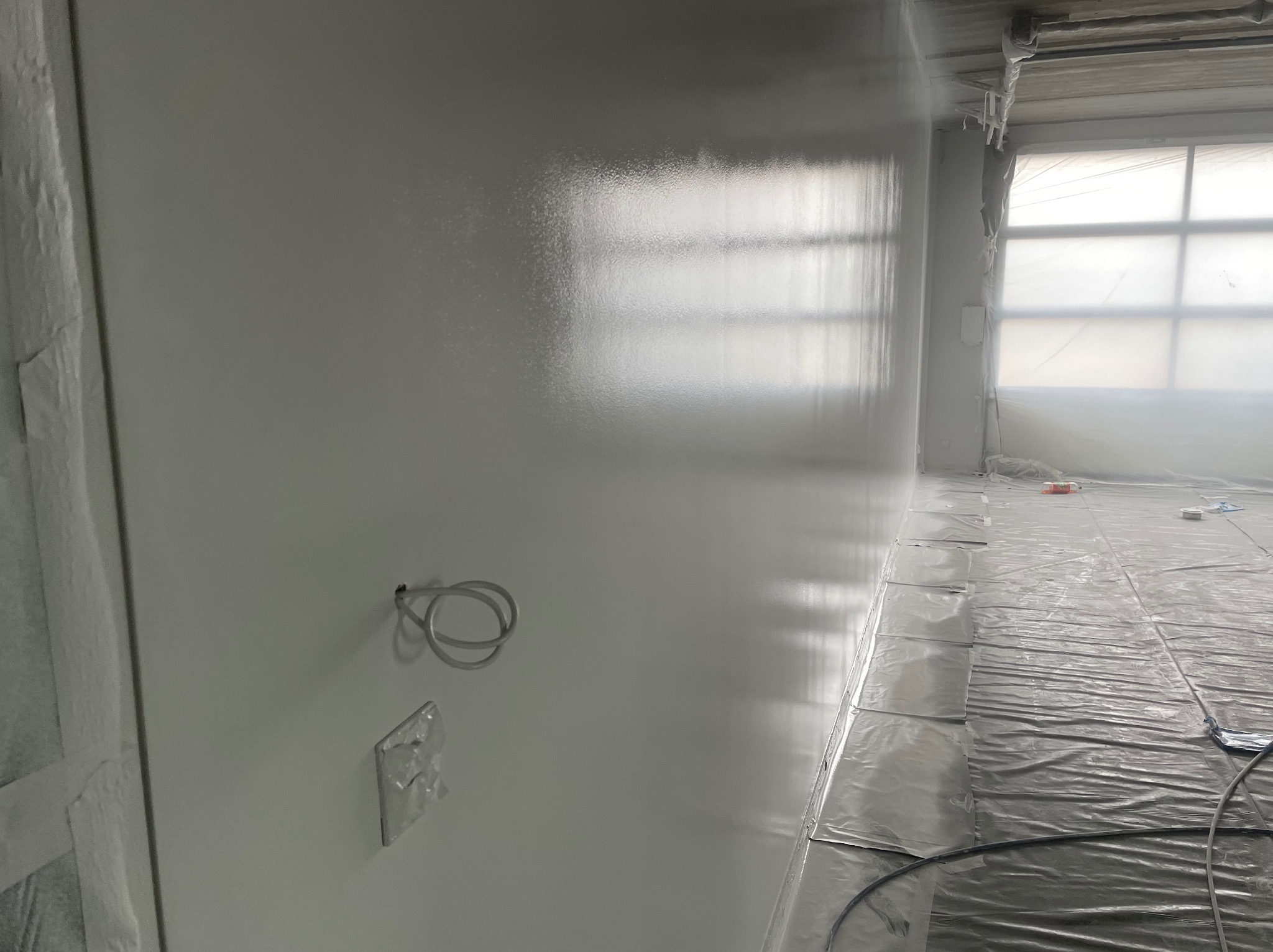 Świeżo pomalowane na biało ściany wnętrza z widocznymi zabezpieczeniami folią malarską na podłodze i oknach. Widoczne gniazdko elektryczne i wystające przewody.