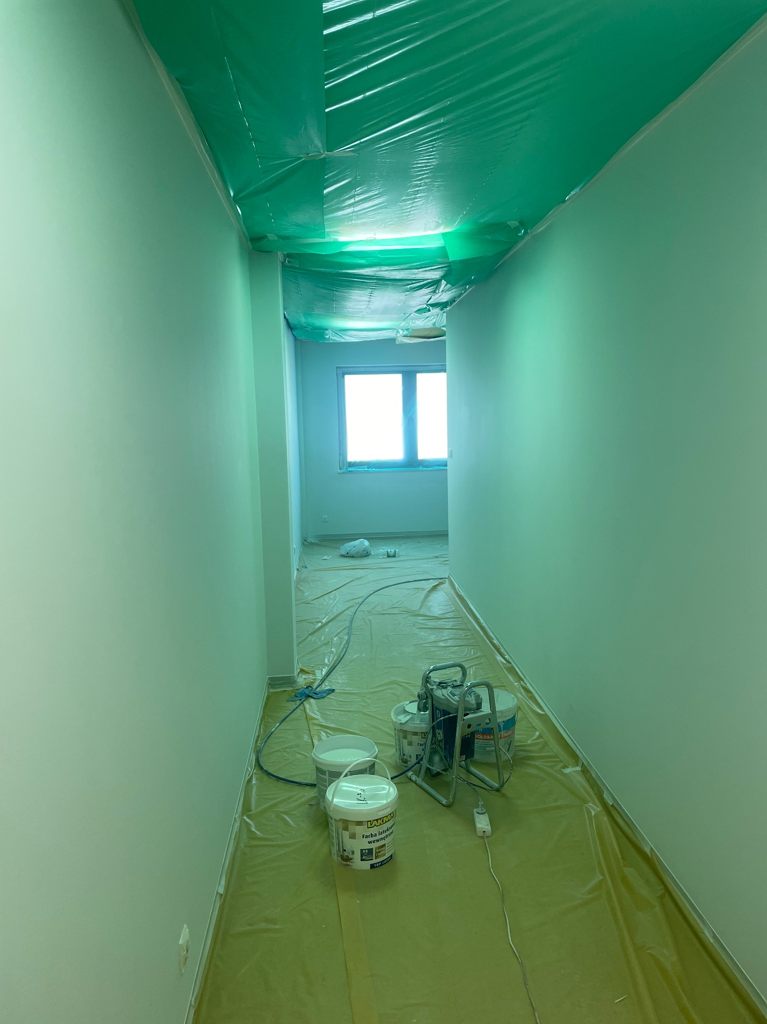 Korytarz w trakcie malowania natryskowego na jasnozielono, z zabezpieczoną folią podłogą i sufitem, widoczne wiadra z farbą i urządzenie malarskie.