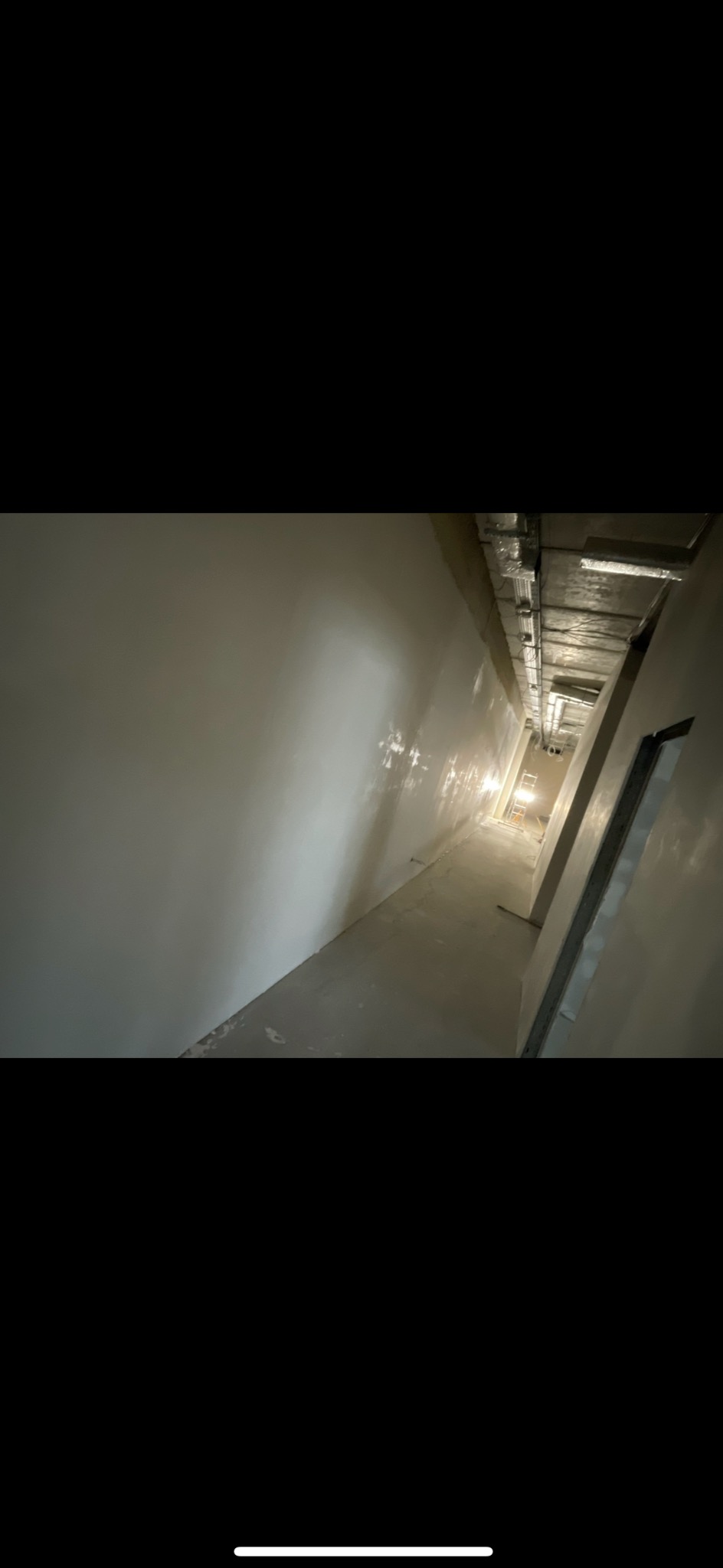 Długi, wąski korytarz po szpachlowaniu ścian i podłogi, z widocznymi instalacjami pod sufitem. W tle drabina i światło wpadające z końca korytarza.