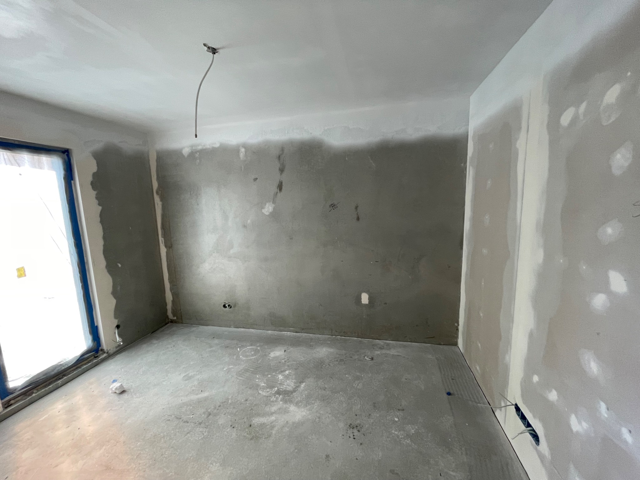 Pokój w trakcie szpachlowania: ściany częściowo pokryte gładzią, widoczne kable, okno zabezpieczone folią. Podłoga betonowa, sufit biały z wystającym kablem.