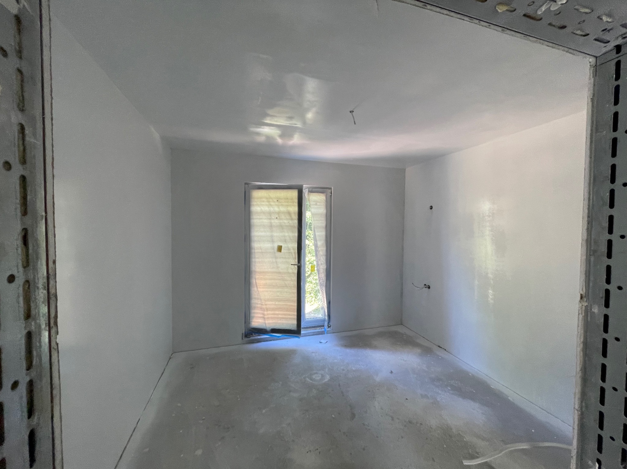 Pokój po szpachlowaniu, przygotowany do malowania. Widoczne gładkie ściany, okno zabezpieczone folią i metalowe profile w rogach pomieszczenia.