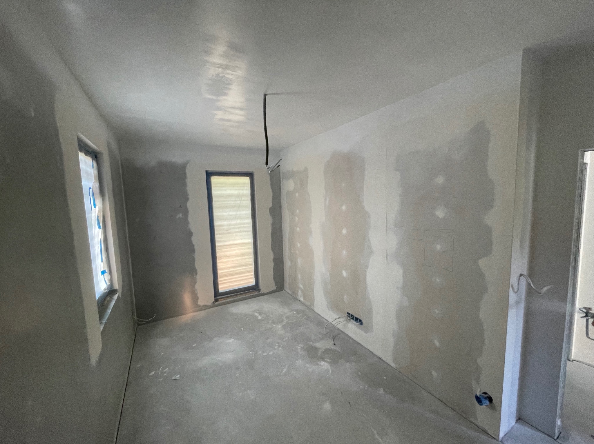 Pokój w trakcie szpachlowania, widoczne gładzie na ścianach, okno zabezpieczone folią, wystające kable elektryczne. Ściany przygotowane pod malowanie.